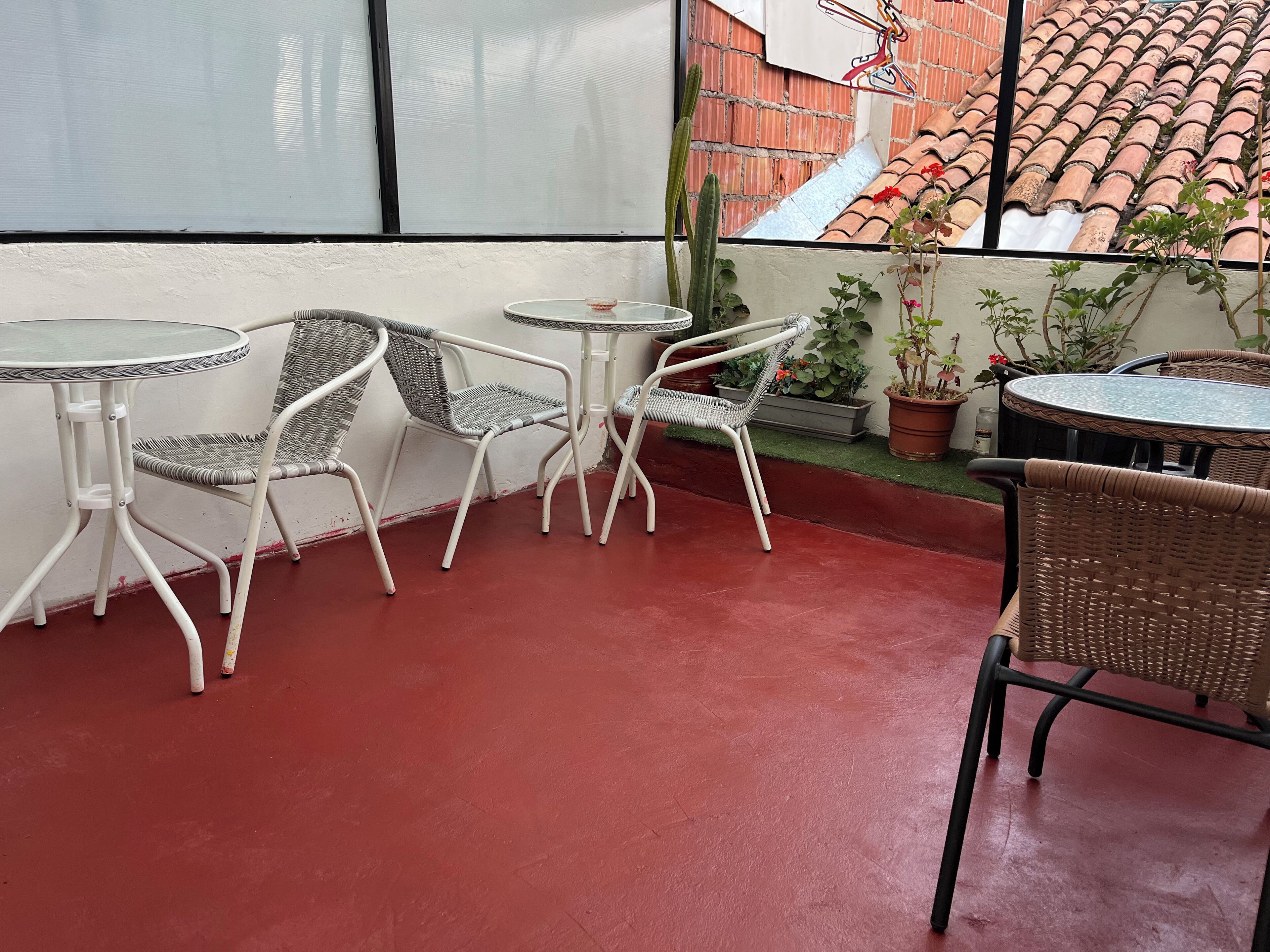 Una terraza con sillas y mesas de mimbre blanco, piso rojo y plantas en macetas.
