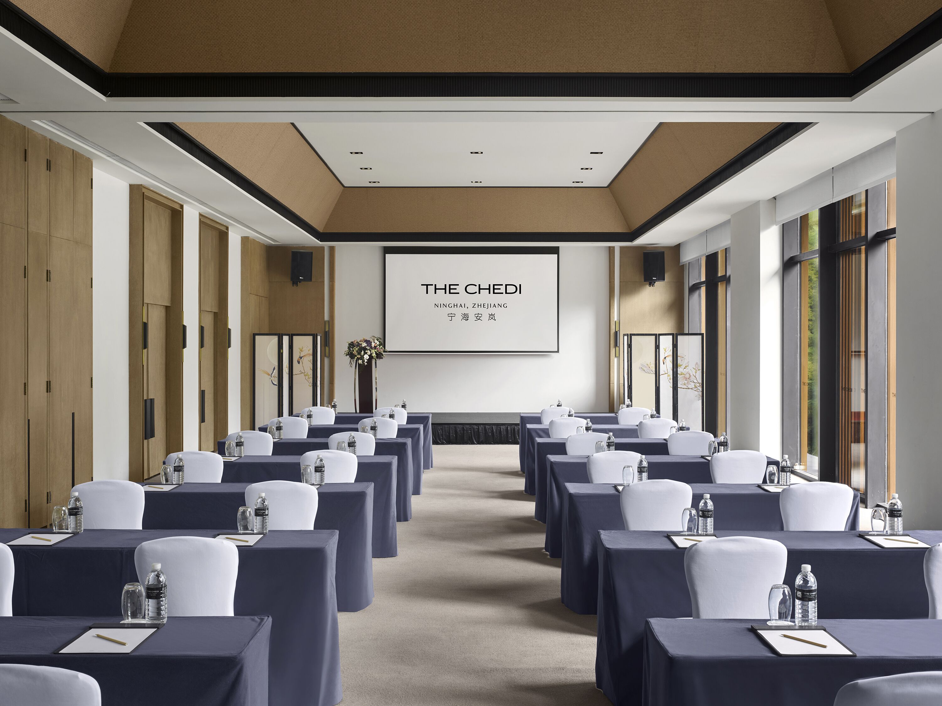 Ein Konferenzraum mit Tischen und Stühlen, einer Projektionsleinwand, auf der 'THE CHEDI NINGHAI, ZHEJIANG' zu sehen ist, und einem großen Fenster auf der rechten Seite.