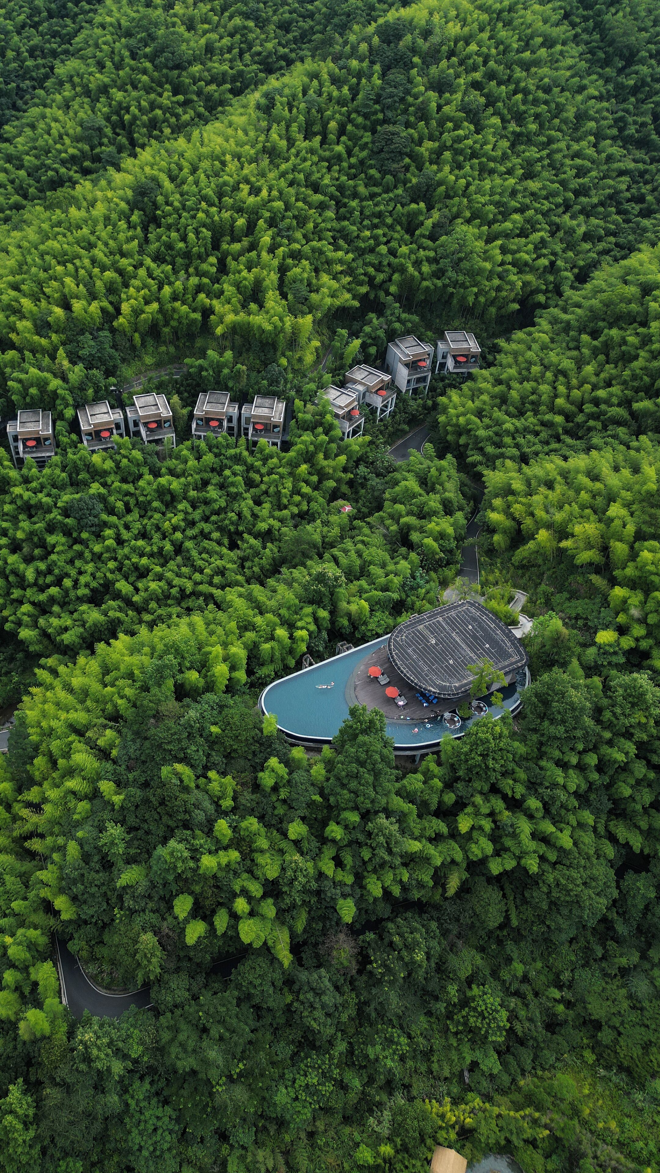 An aerial view of a resort with multiple modern buildings surrounded by dense forest. There is a large circular pool area with a covered seating area.