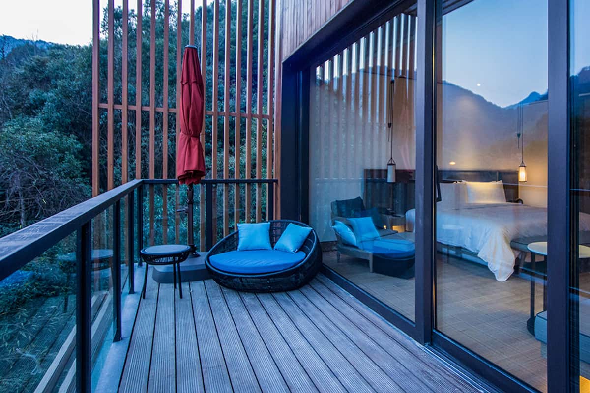 Eine moderne Außenterrasse mit Holzboden, ein glaskonstruierter Schlafbereich mit Bett und Nachttisch sowie ein roter Sonnenschirm.