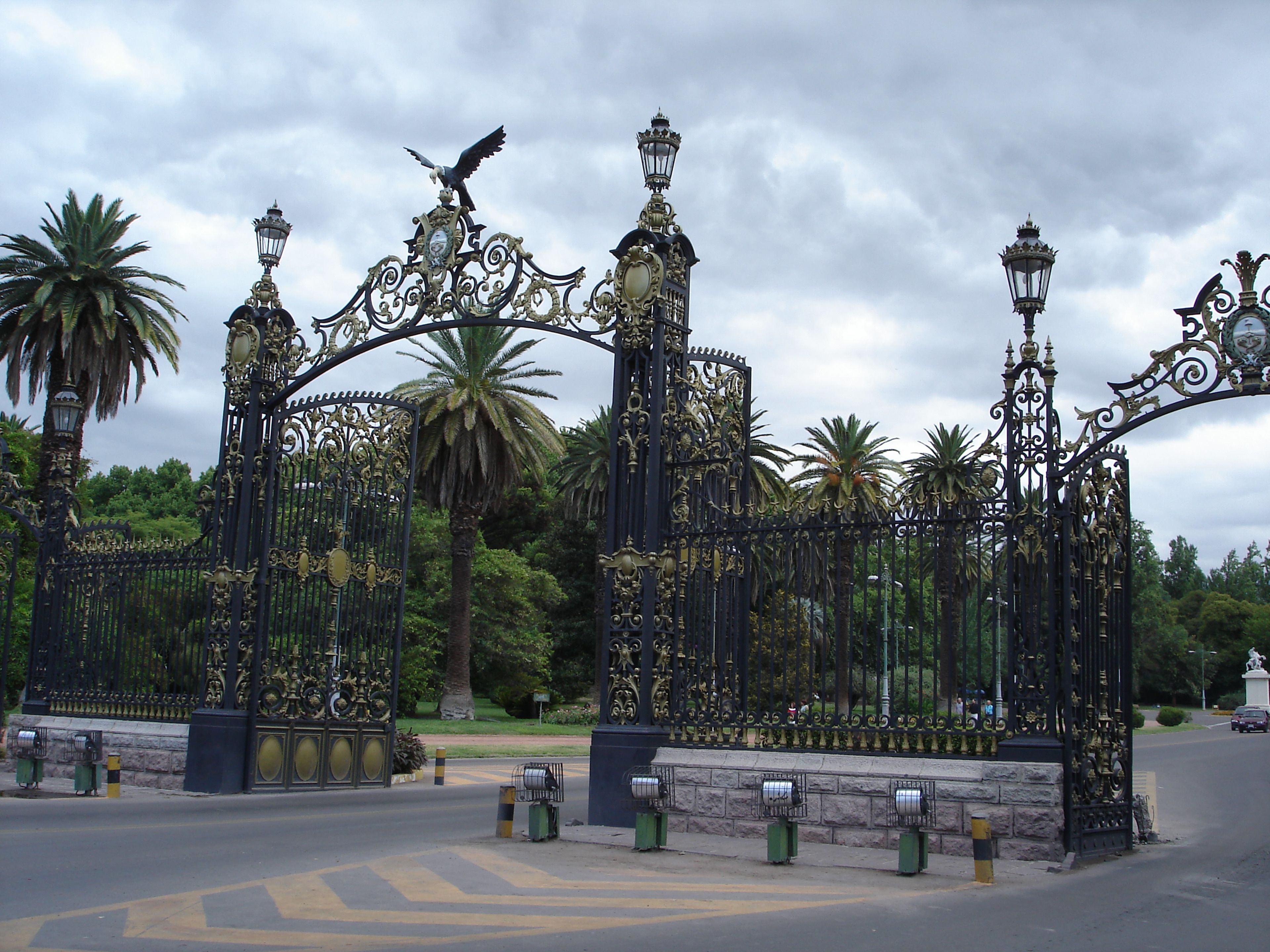 Una reja de hierro forjado con diseños intrincados y una escultura de un pájaro, rodeada de palmeras y follaje.