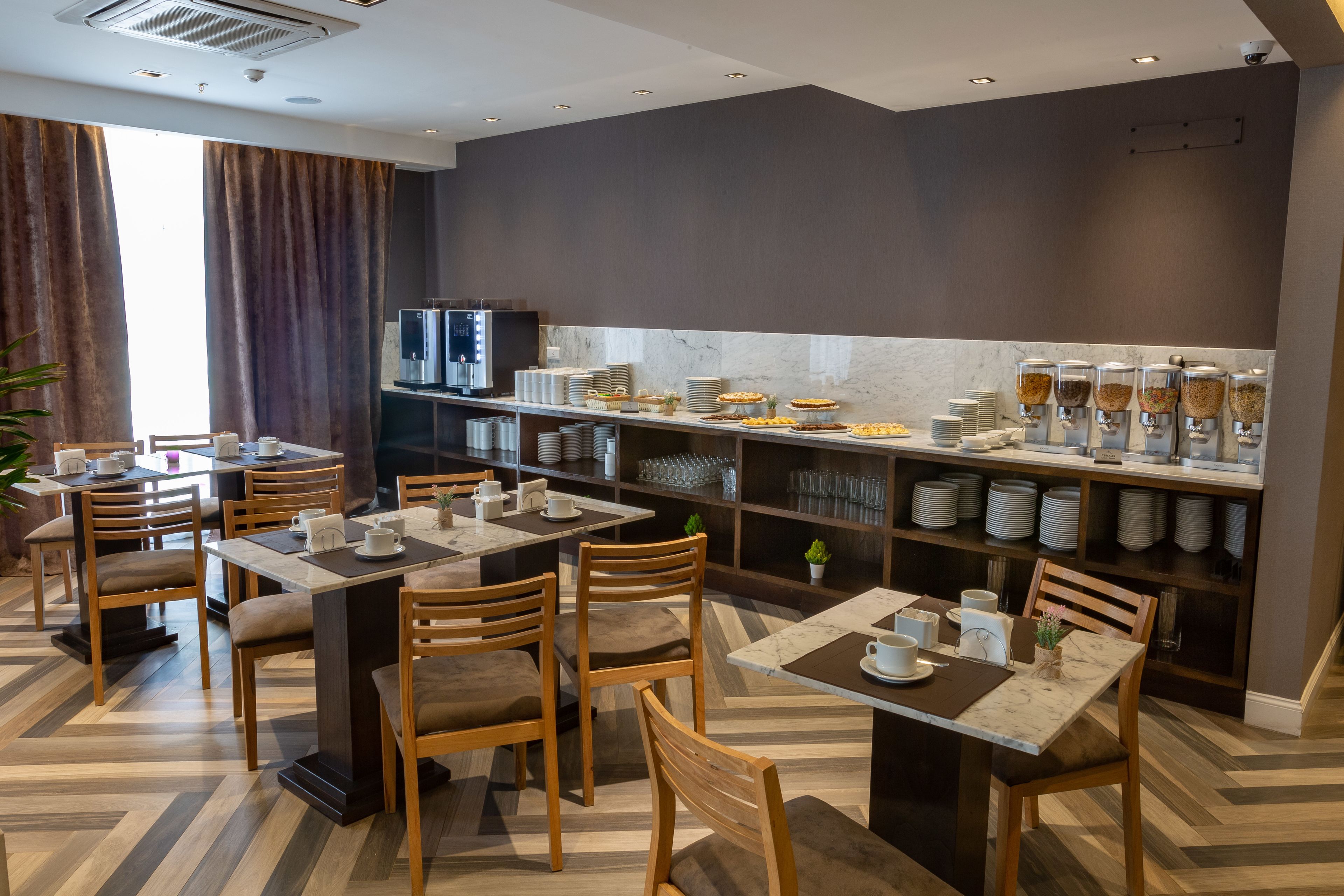 A modern dining area with wooden tables and chairs, a buffet with various food items, and a kitchen area with a refrigerator and shelves.