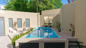 Un área de piscina exterior moderna con una piscina azul cristalina, rodeada de paredes color beige y sillas reclinables blancas.