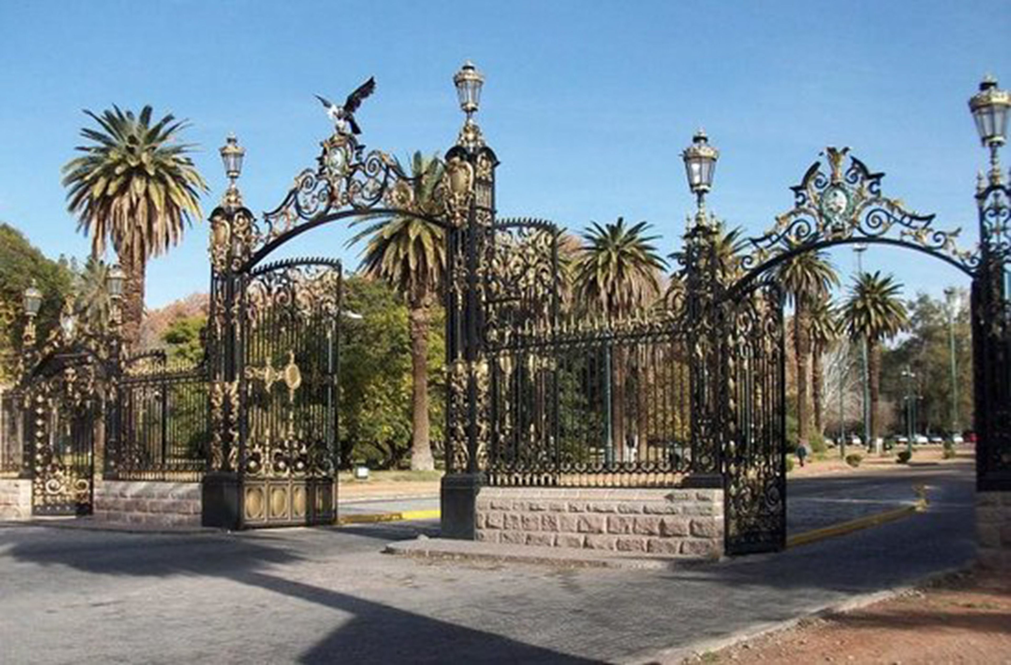 A wrought iron gate with intricate designs and a stone base, surrounded by palm trees and a clear sky.