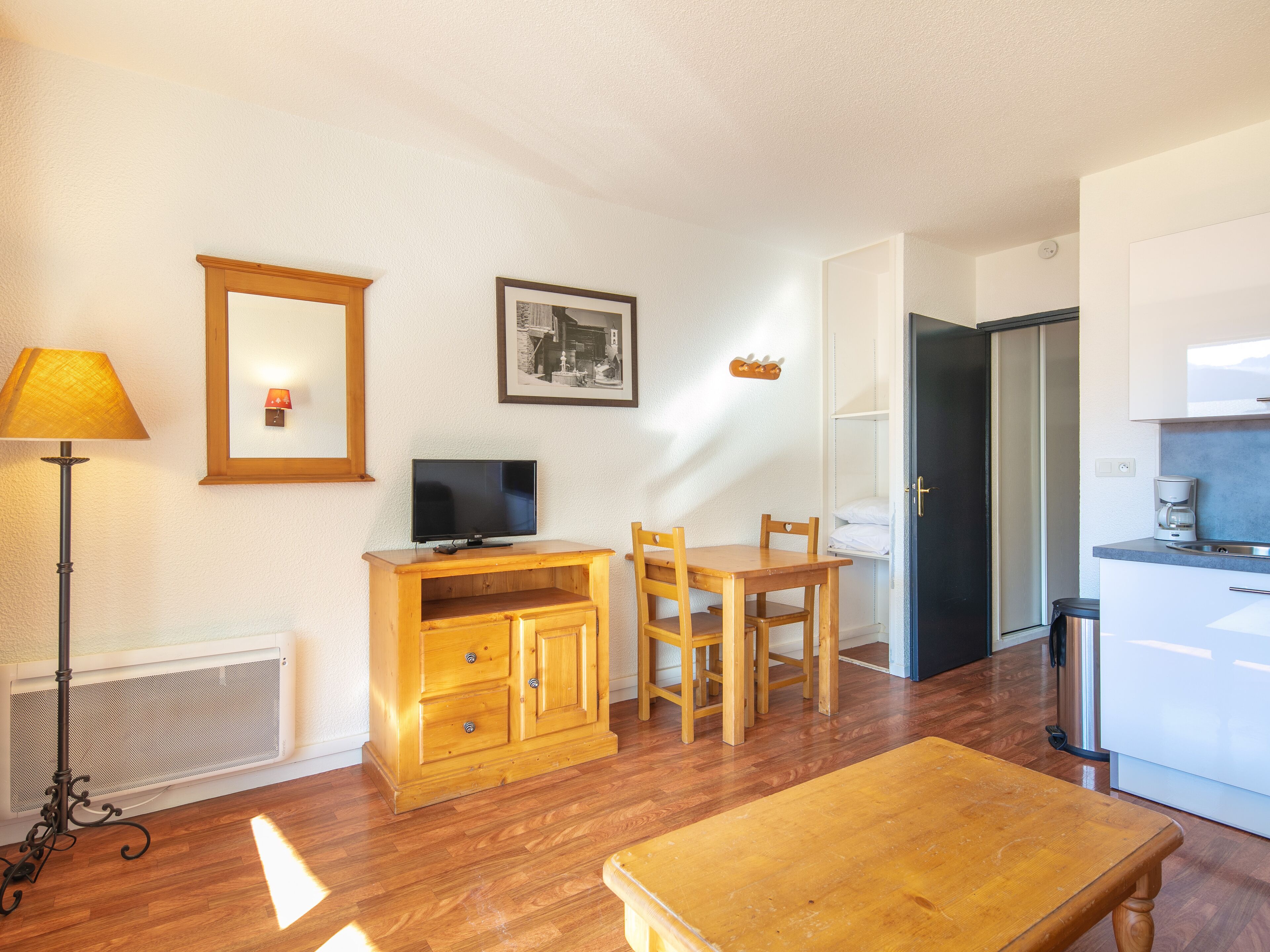 Un espace de vie compact comprenant une table en bois, un téléviseur posé sur un meuble en bois, un tableau encadré au mur et un coin cuisine avec un réfrigérateur et une machine à café.