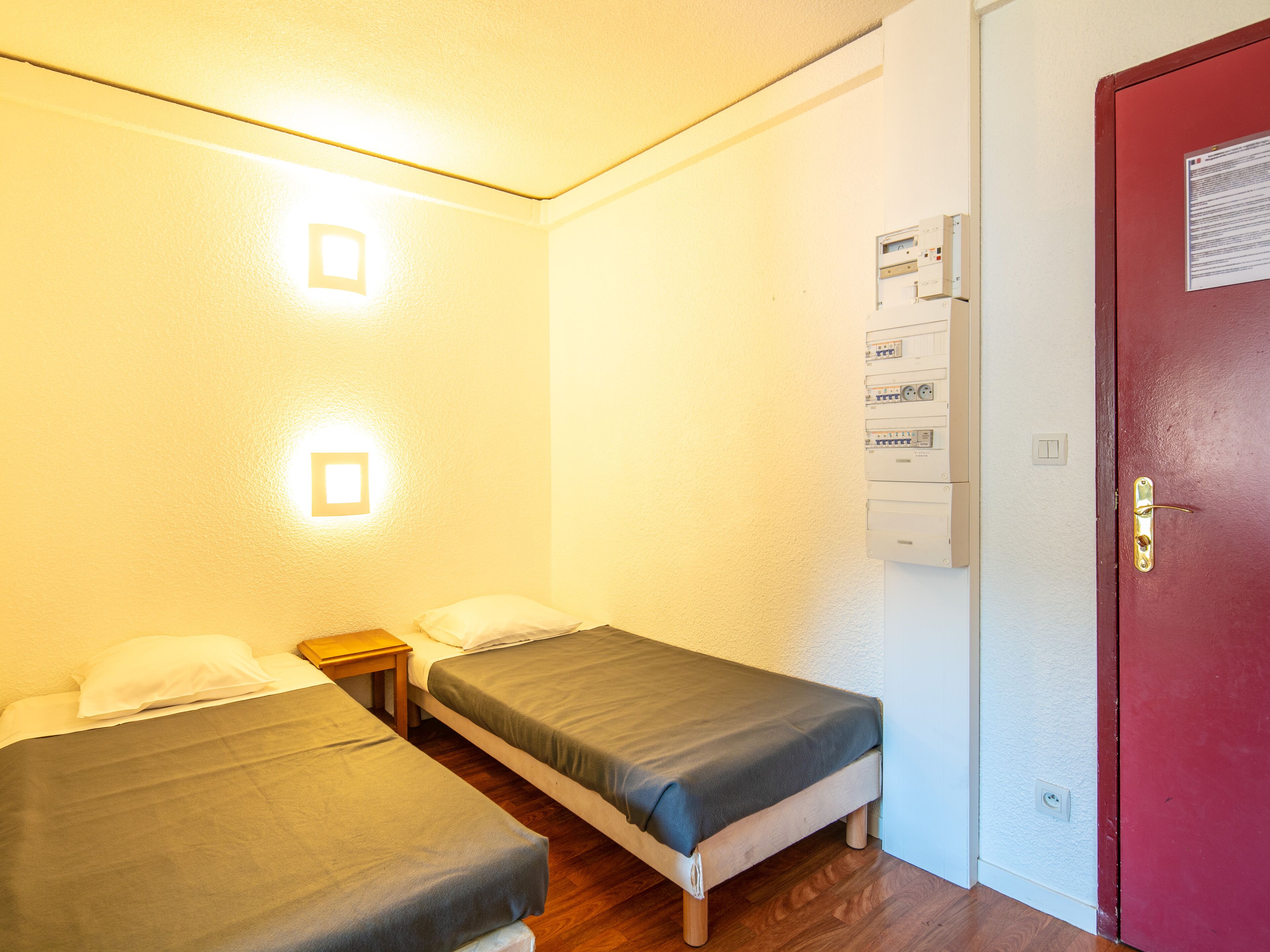 A small, well-lit room with two single beds, a wooden floor, and a red door.