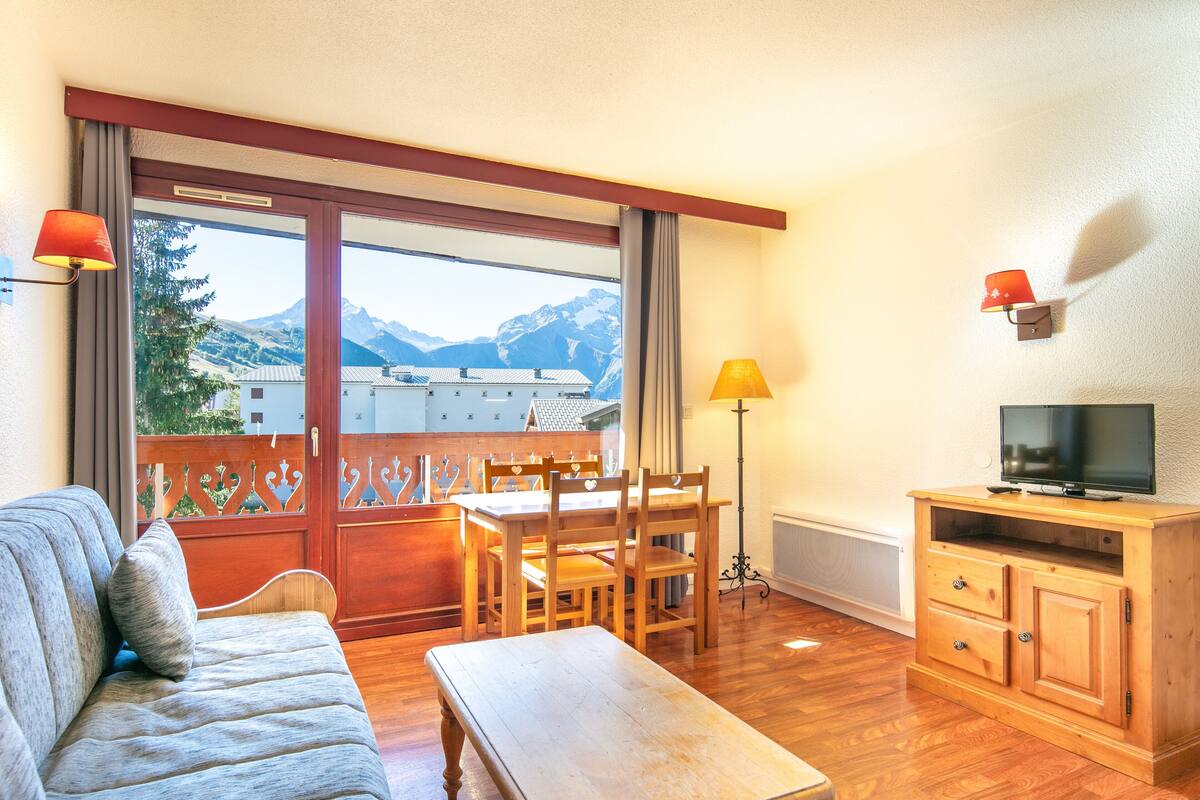 Un salon chaleureux avec un canapé, une table et des chaises en bois, un téléviseur posé sur un meuble en bois, et une vue sur les montagnes par une grande fenêtre.