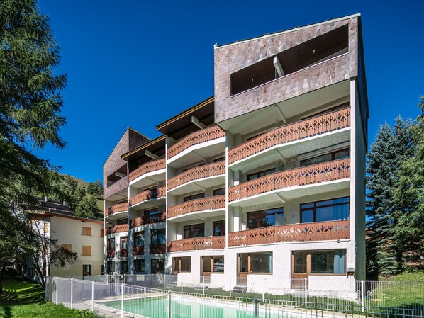 Un hotel de varios pisos con balcones, piscina y montañas al fondo.