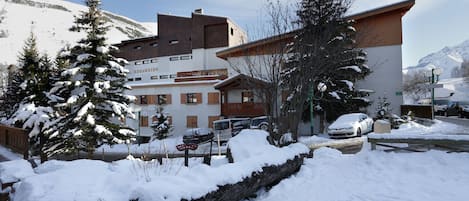 A hotel with a snow-covered landscape, a mountain in the background, and a parking area with cars.