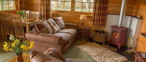 A cozy log cabin interior with a leather sofa, a dining table with chairs, a fireplace, and a window with curtains.