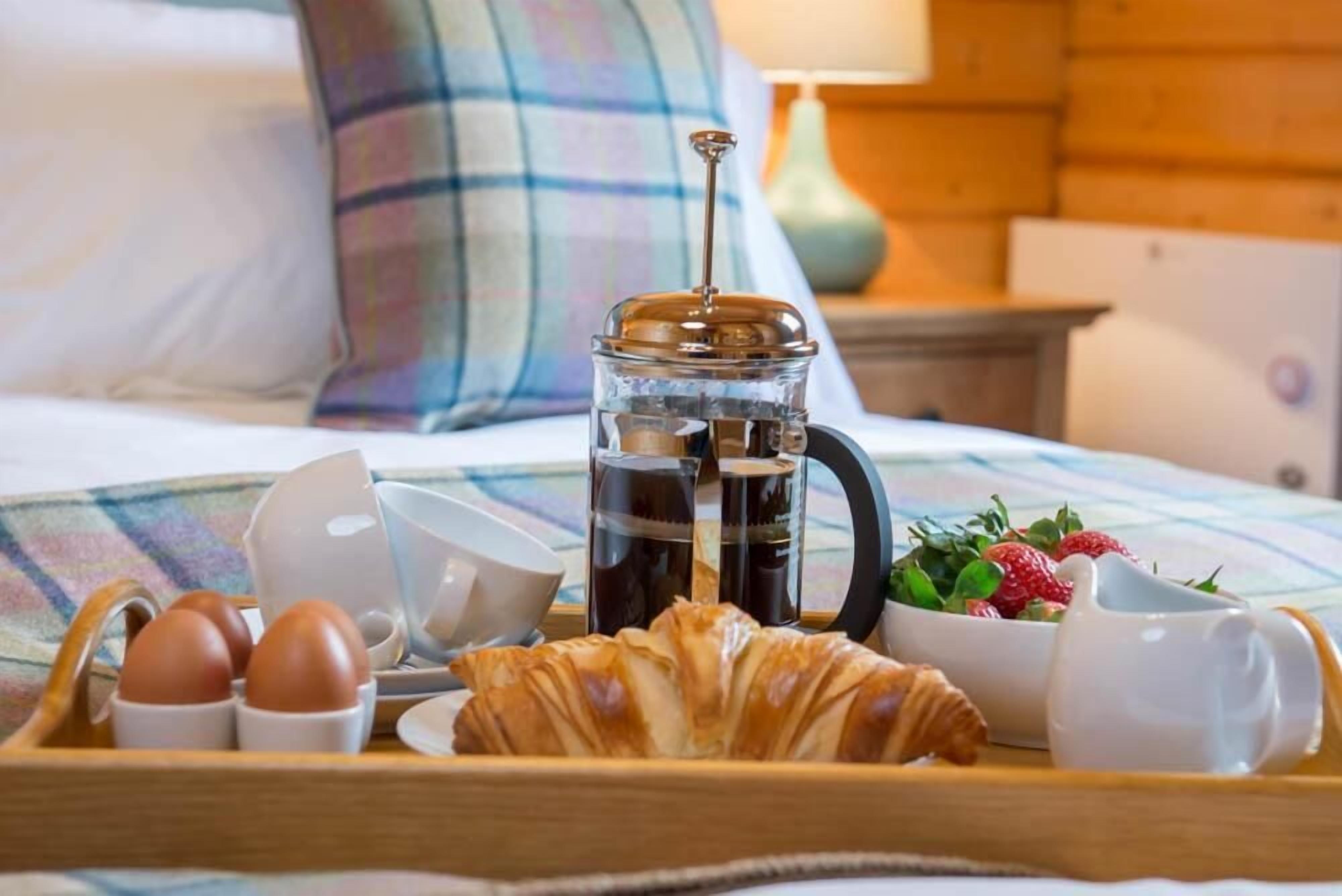 Uma bandeja de café da manhã com ovos, um croissant, café e uma tigela de morangos sobre uma cama com travesseiro xadrez e uma luminária ao fundo.