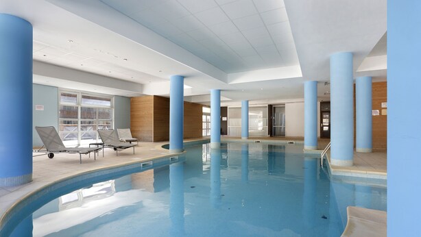 An indoor swimming pool with blue pillars, lounge chairs, and a view of the outside through large windows.