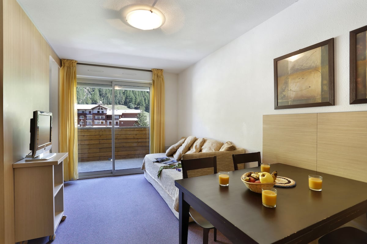 A hotel room with a bed, a dining table with fruit and juice, a TV, and a balcony with curtains.