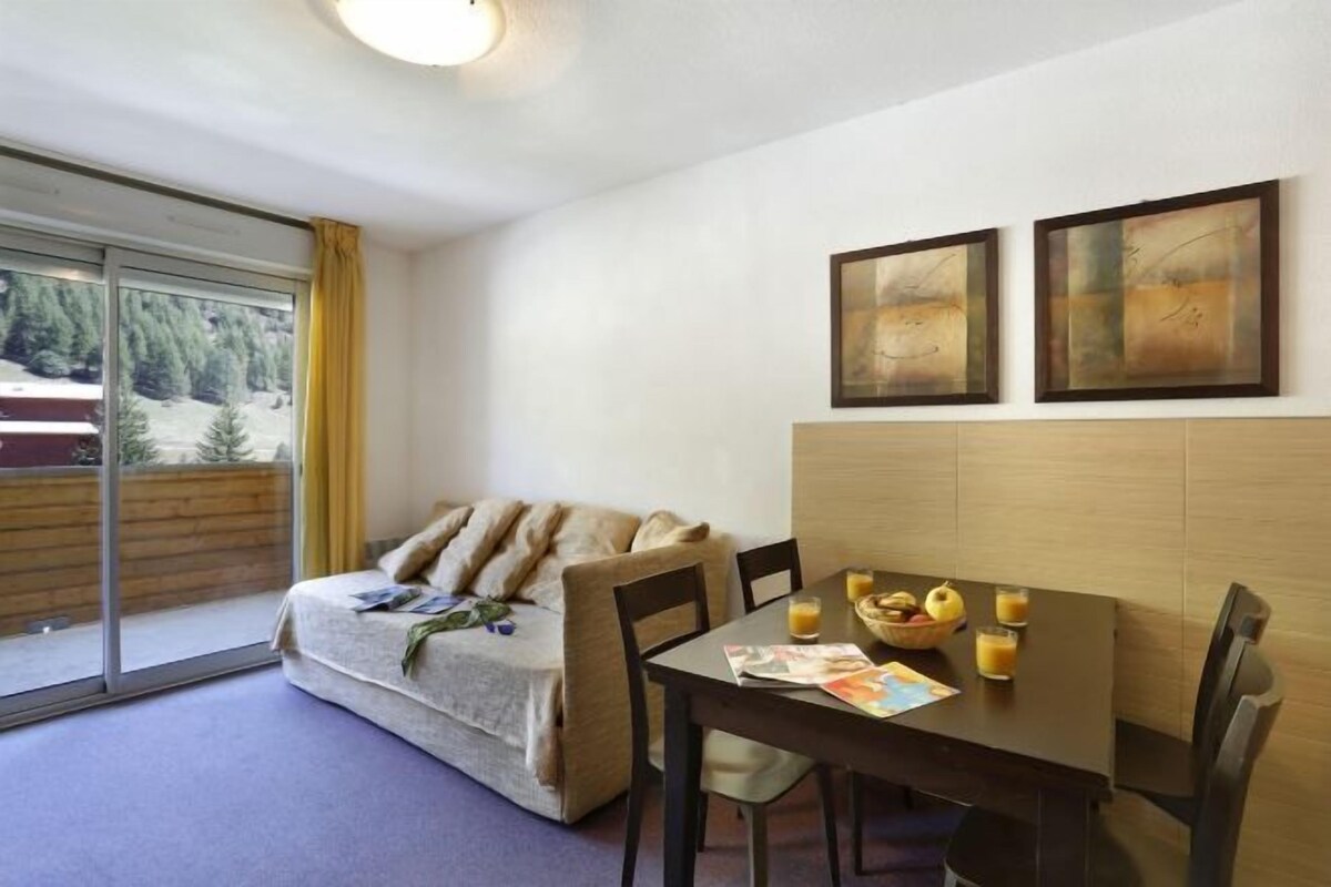 A cozy room with a sofa, a dining table with chairs, and a window with a view of trees.