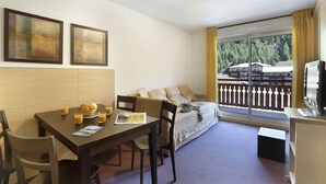A room with a dining table, chairs, a sofa, a TV, and a window with a view of a building and trees.