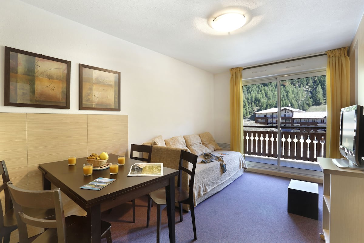 A room with a dining table, chairs, a sofa, a TV, and a window with a view of a building and trees.