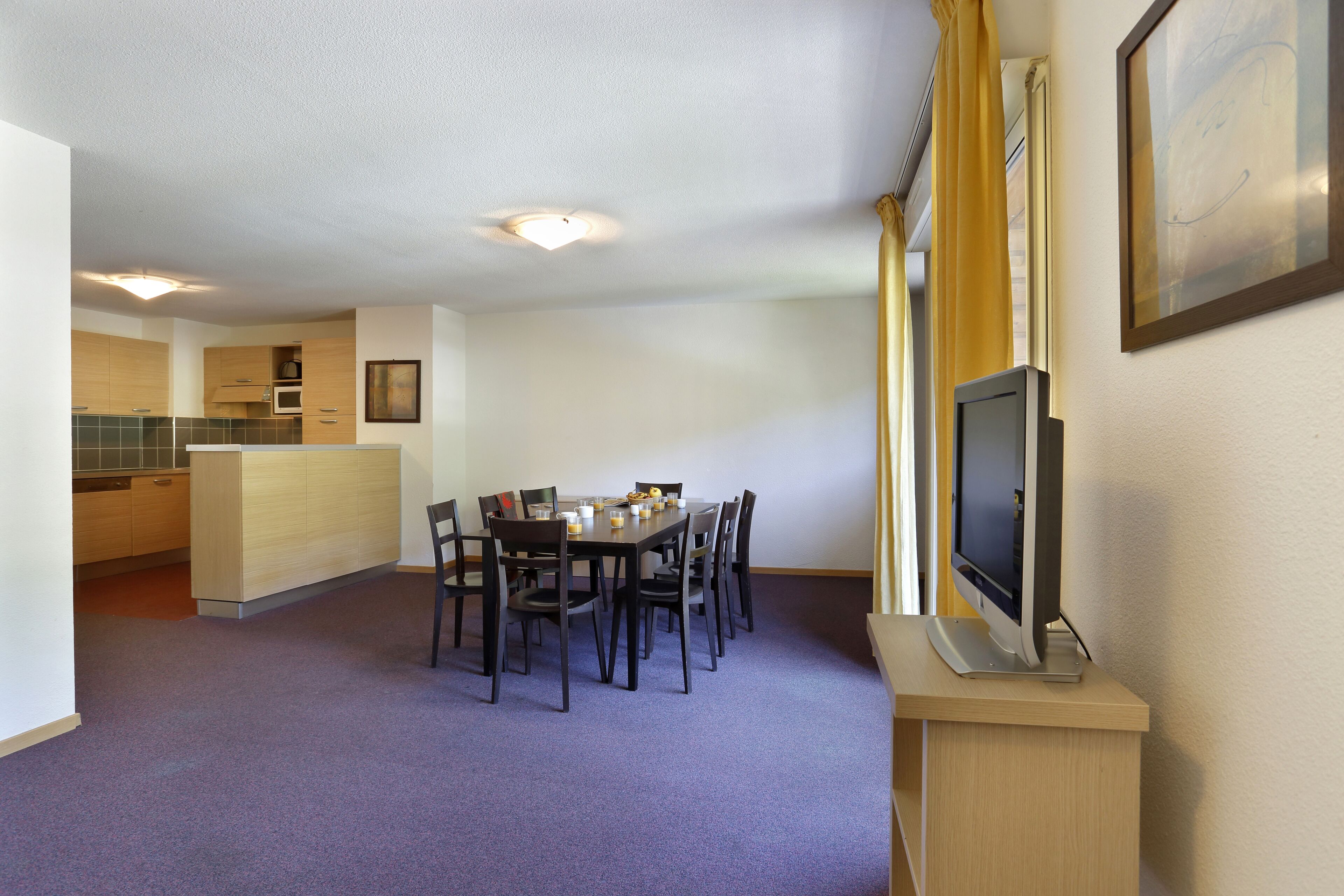 A dining area with a table and chairs, a television on a stand, and a kitchen in the background.
