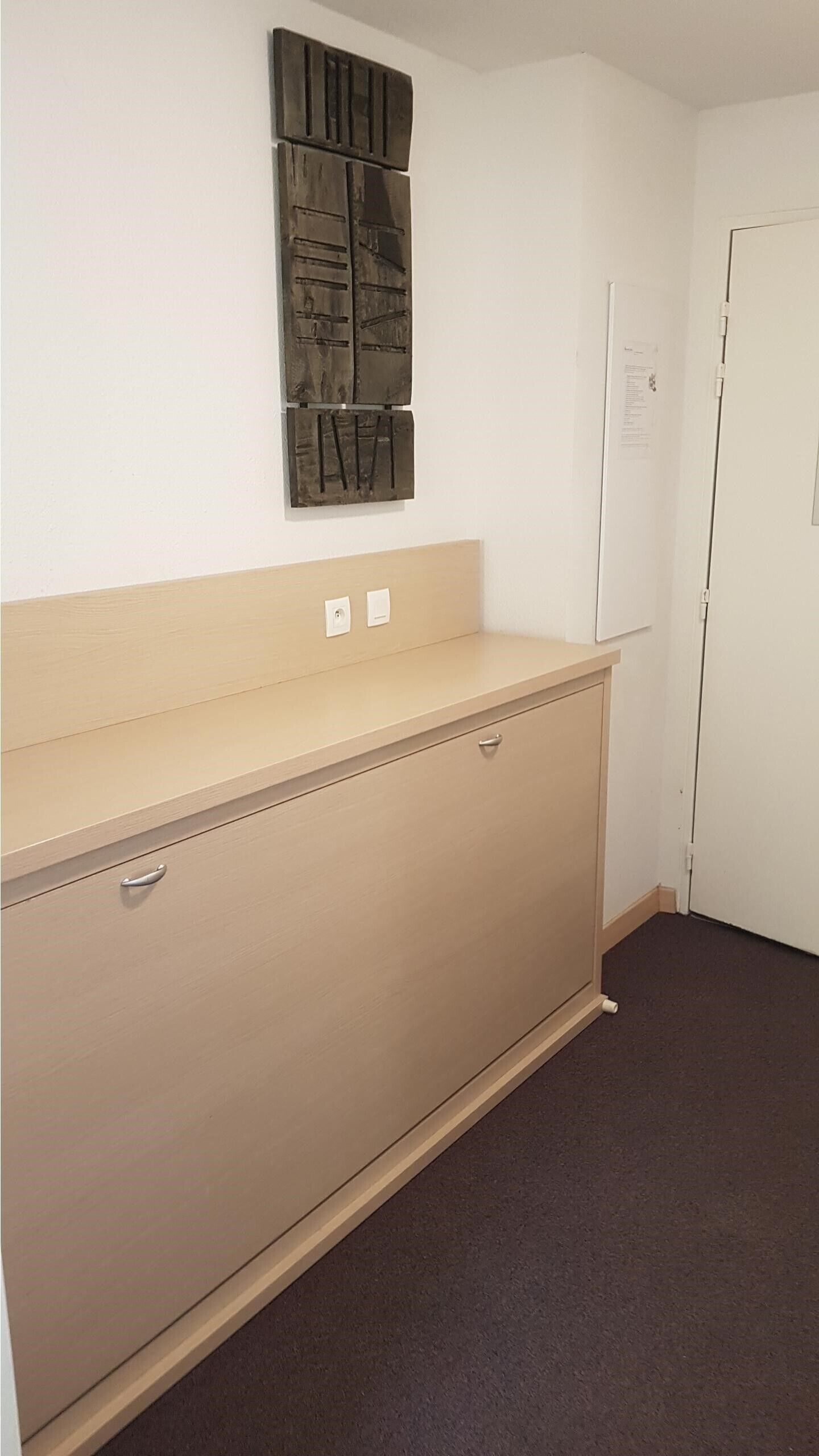 A room with a beige dresser, a small table, and a wall-mounted artwork. There is a door with a sign on it.