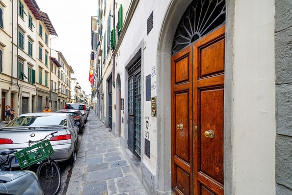 Una calle estrecha con coches aparcados y una bicicleta, una puerta de madera con tirador de latón y edificios con balcones y contraventanas.