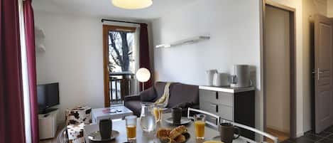 A modern living room with a sofa, a coffee table, and a TV. There is a dining area with a table set for breakfast, including fruits, pastries, and drinks.