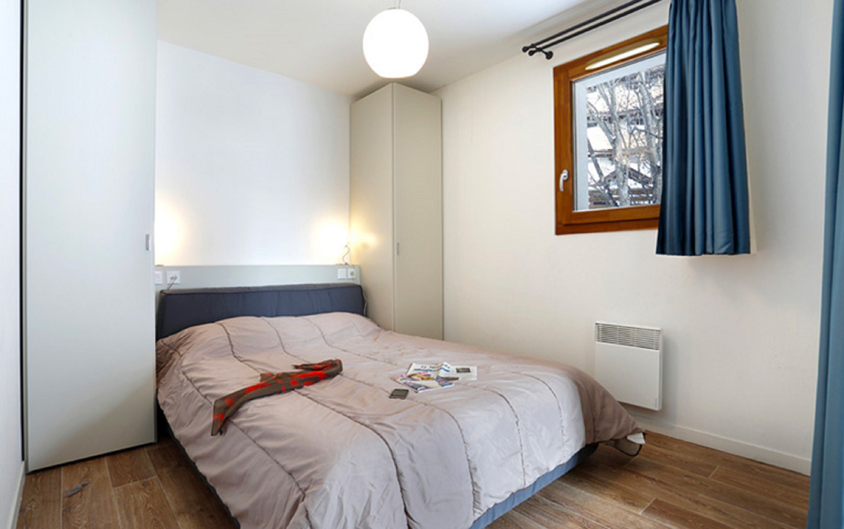 A bedroom with a bed, a window with curtains, a radiator, and a wall-mounted light fixture.