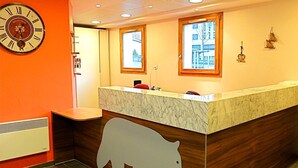 A hotel reception area with a marble counter, wooden paneling, a bear silhouette, a clock, and windows with city views. - Vacanceole - Au Coeur des Ours (Les Deux Alpes)