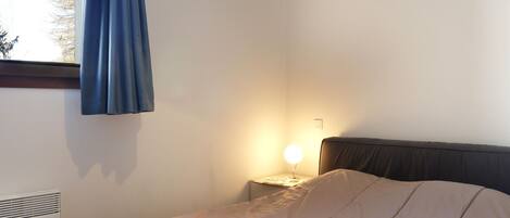 Ein Schlafzimmer mit einem Bett, einer Nachttischlampe, einem Fenster mit Vorhängen und einer Deckenleuchte.