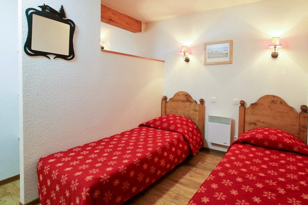Ein Zimmer mit zwei Holzbetten, beide mit einer roten Bettdecke mit Schneeflockenmuster. Zwei Wandlampen, ein gerahmtes Bild an der Wand und ein kleines Spiegel links sind vorhanden.