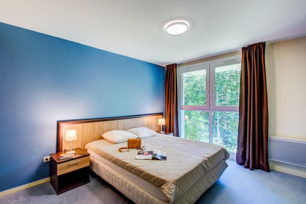 Ein Hotelzimmer mit Bett, Nachttischen, einem Fenster mit Vorhängen und Blick auf Bäume.