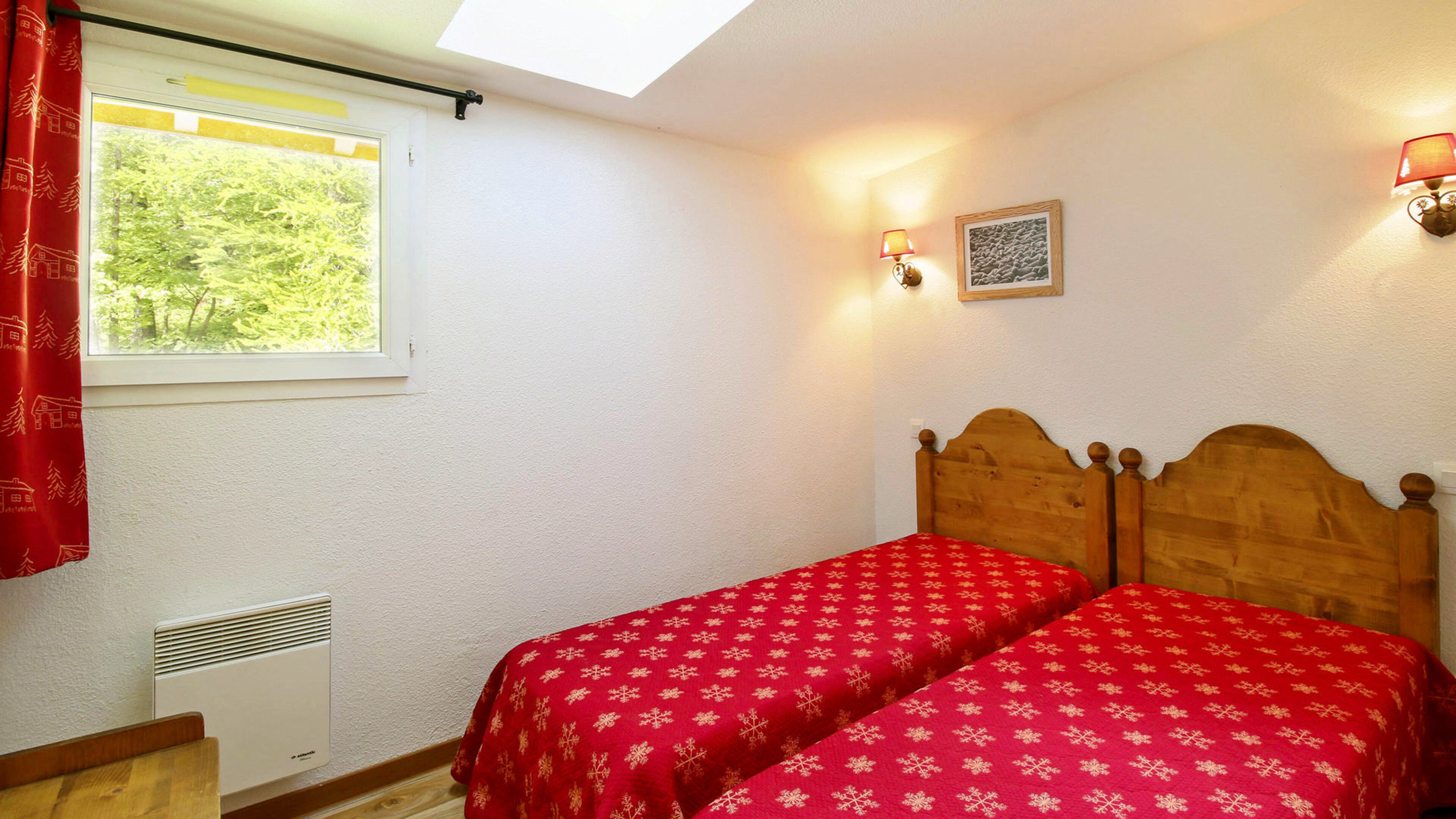 A bedroom with two single beds, a window with a view of trees, a radiator, and a wall-mounted lamp.