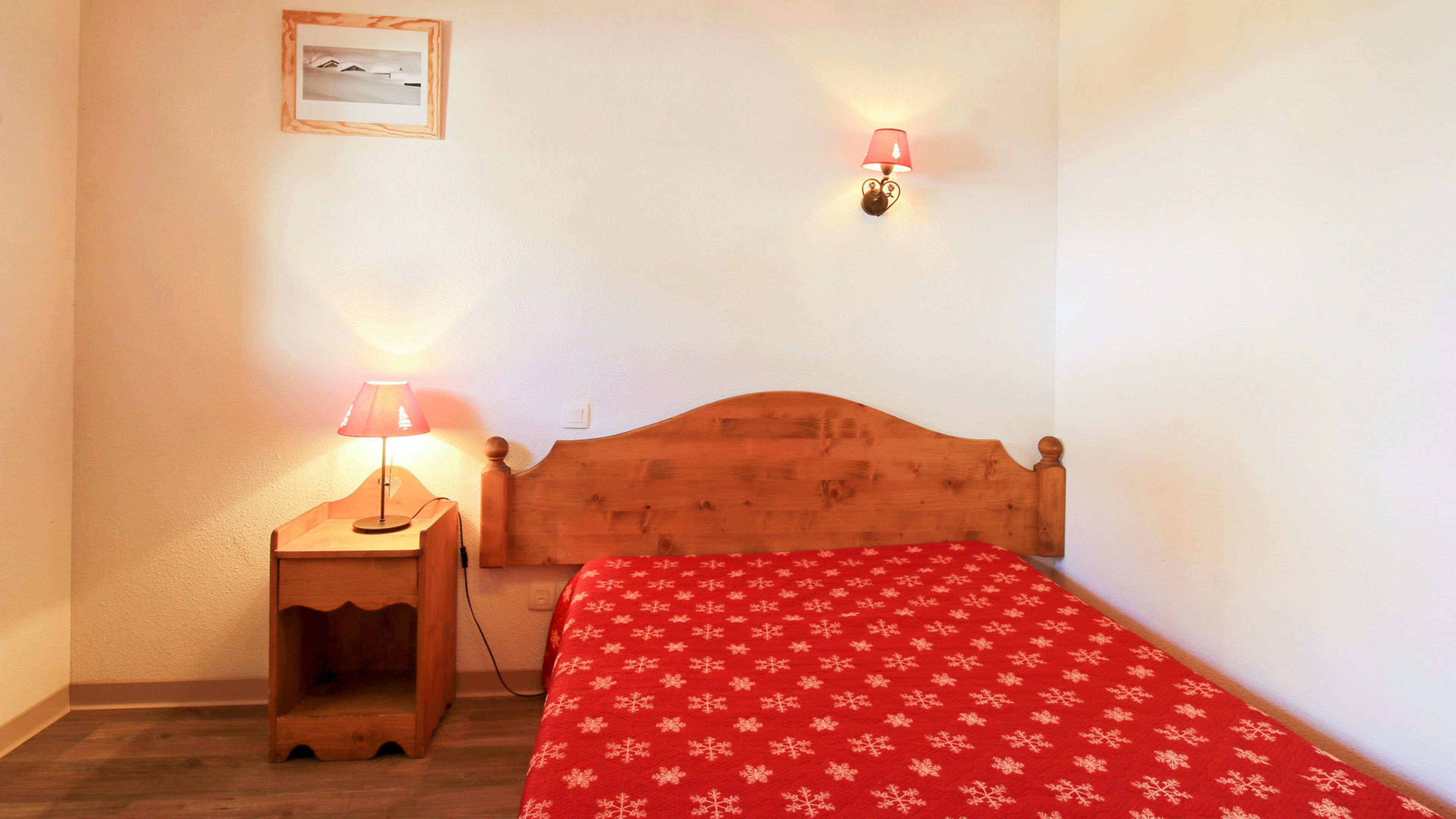 Ein kleines Schlafzimmer mit einem hölzernen Bett, einem Nachttisch mit Lampe, einer roten Tagesdecke und einem gerahmten Bild an der Wand.