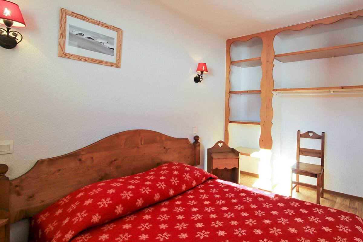 Ein Schlafzimmer mit hölzernem Kopfteil, einem Bett mit roter Decke, einem Nachttisch mit Lampe, einem gerahmten Bild an der Wand und einem Fenster mit Holzläden.