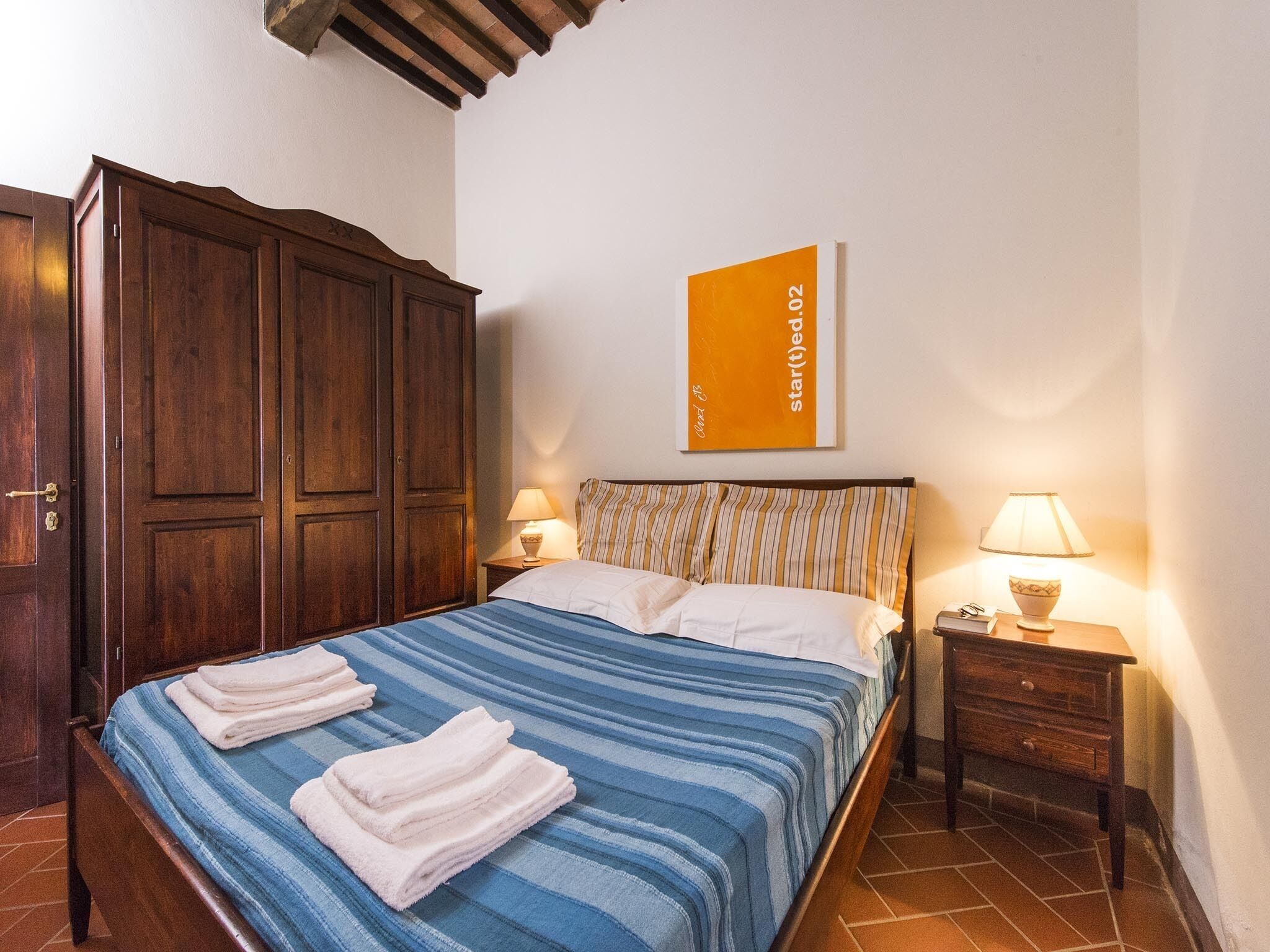 A neatly made bed with striped bedding and two folded towels, a wooden wardrobe, a bedside table with a lamp, and a framed artwork on the wall.