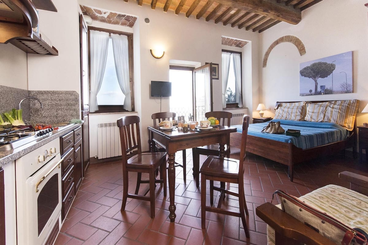 A cozy kitchen with a stove, dining table, and chairs. There is a bed with a blue bedspread and a lamp on a bedside table.