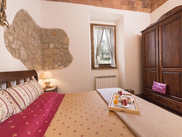 A bedroom with a stone wall, a wooden bed, a nightstand with a lamp, a window with curtains, and a wooden wardrobe. - Country House il Ciliegio (Roccastrada)