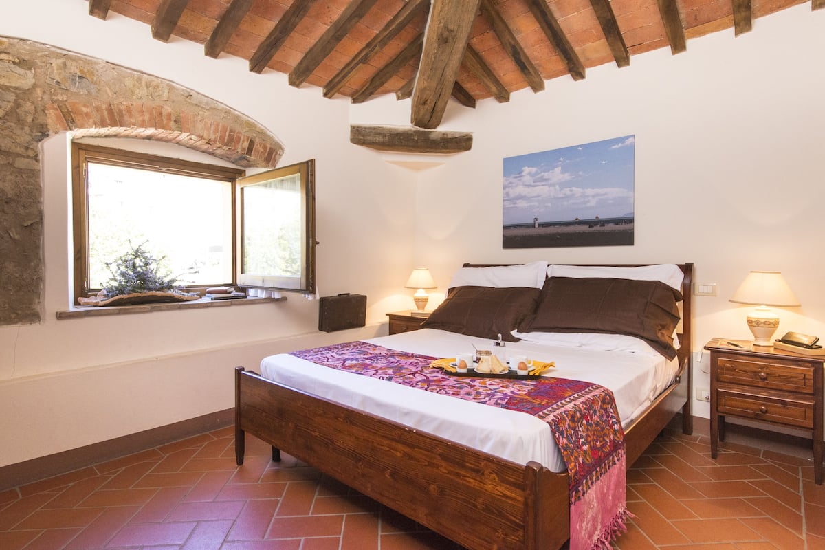A bedroom with a wooden bed, two bedside lamps, a window with a view, and a painting on the wall.