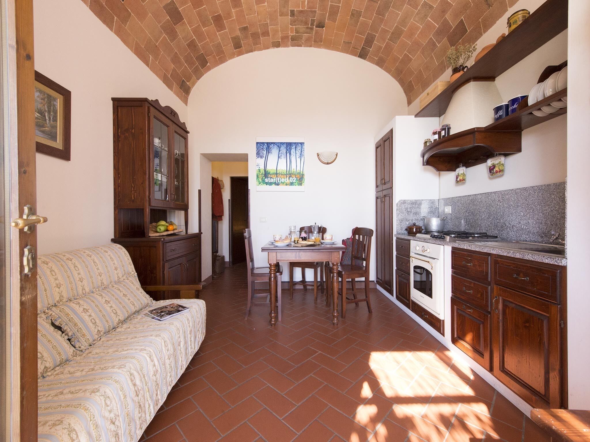 A cozy kitchen with a dining table, chairs, and a sofa. There is a brick ceiling and wooden cabinets.