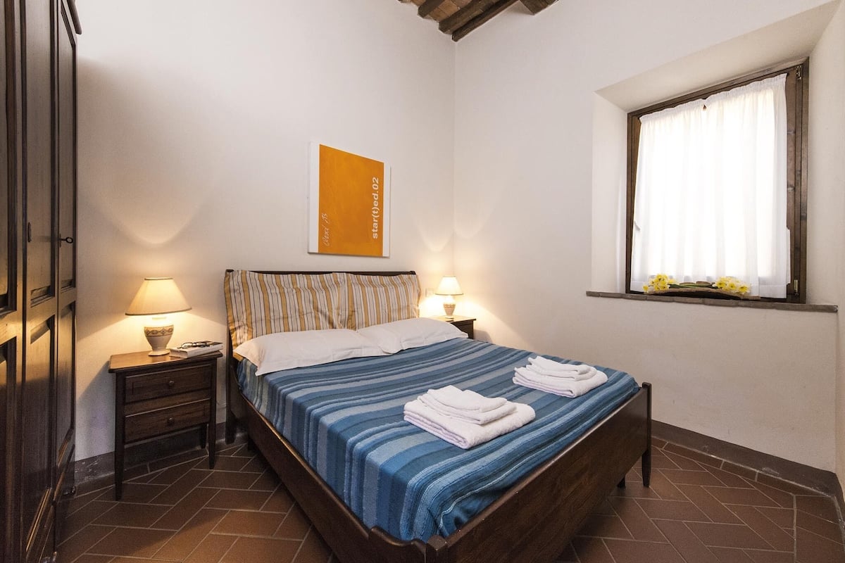 A bedroom with a wooden bed, two bedside tables with lamps, a window with curtains, and a framed picture on the wall.