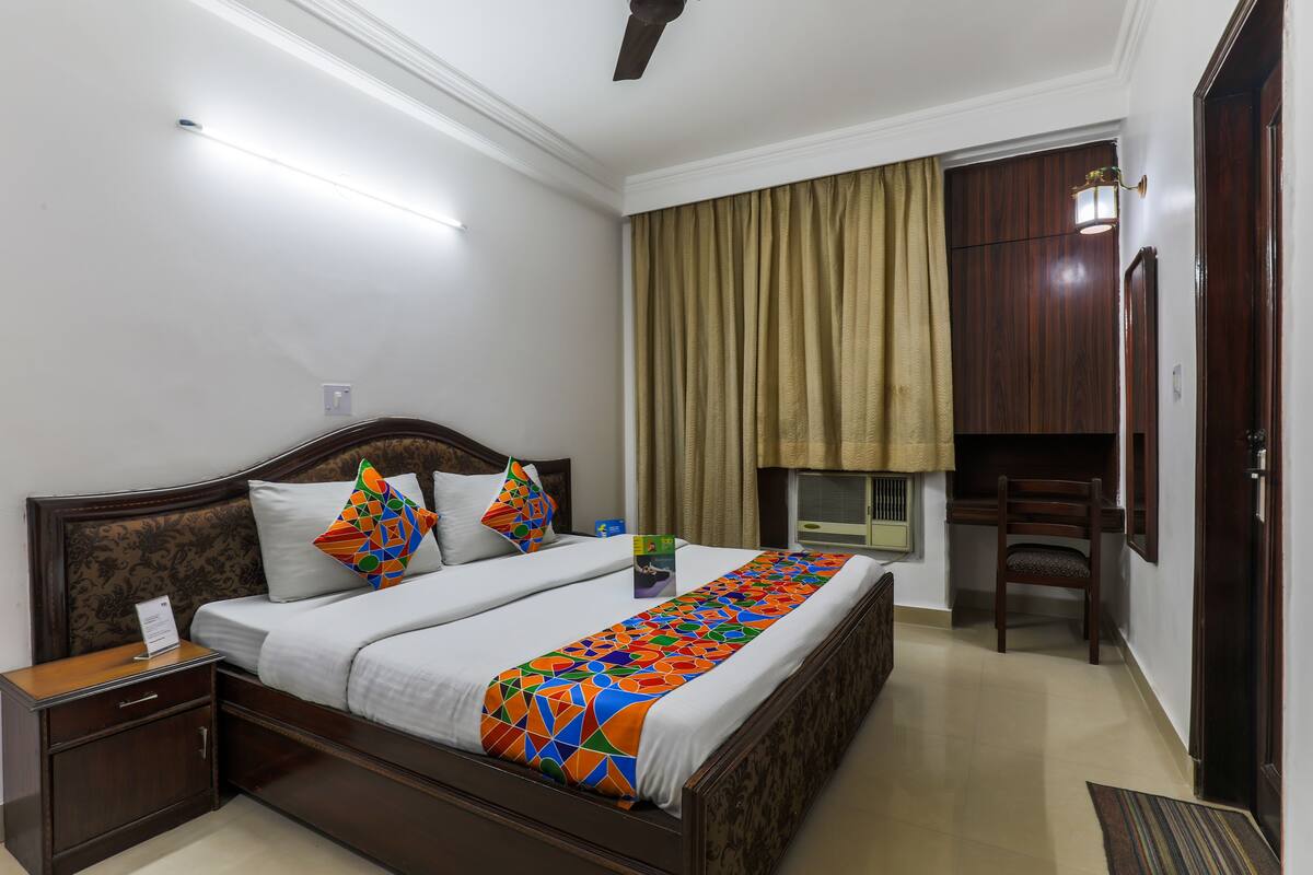 A neatly made bed with a colorful geometric throw, a wooden nightstand, a desk with a chair, and a window with curtains.