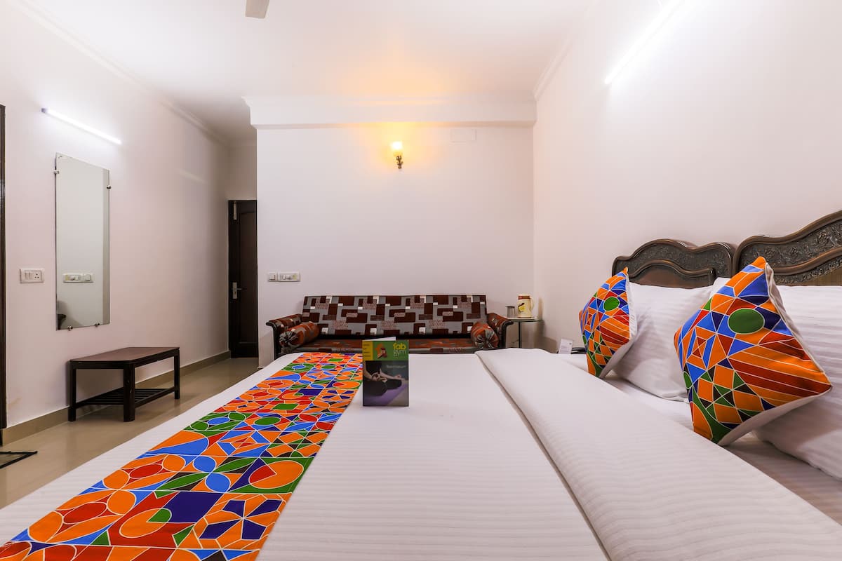 A hotel room with a long, colorful bed runner, a large bed with patterned pillows, a sofa bed, a small table, and a wall-mounted mirror.
