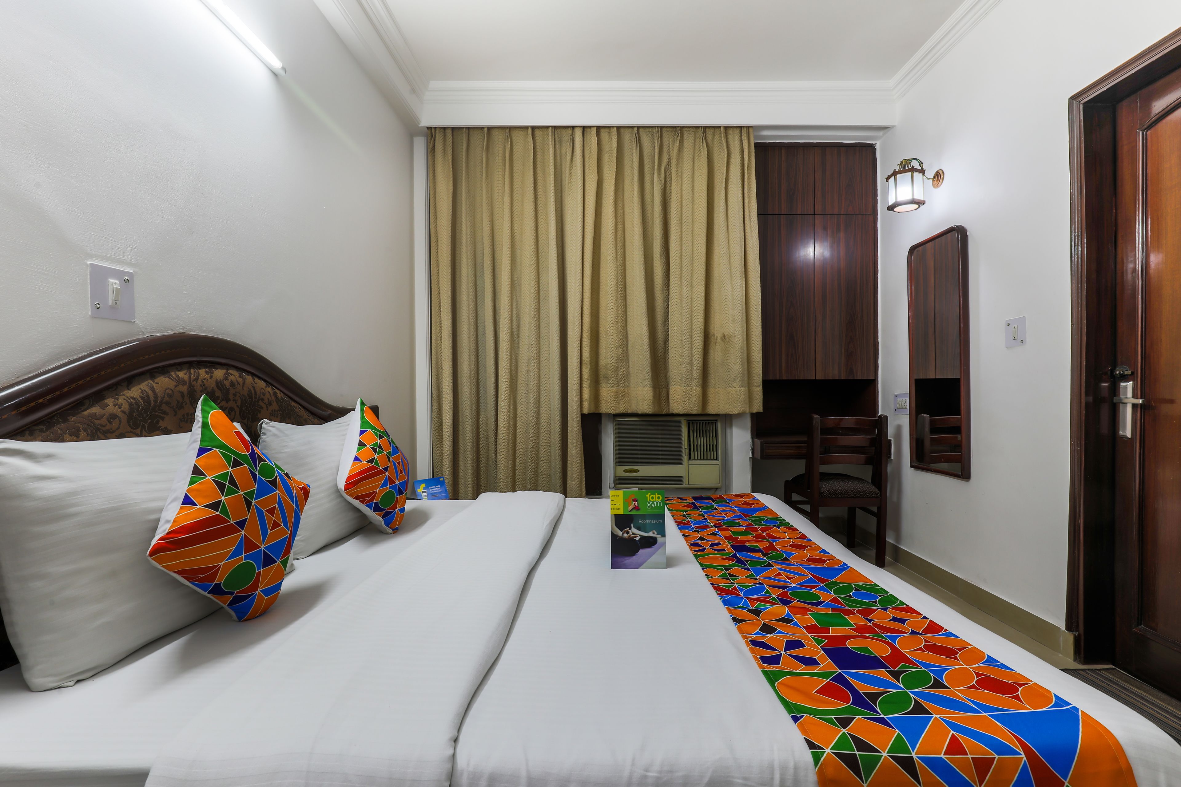 Habitación de hotel con cama, almohadones coloridos, corredor estampado en la cama, ventana con cortinas, puerta y una mesita con lámpara.