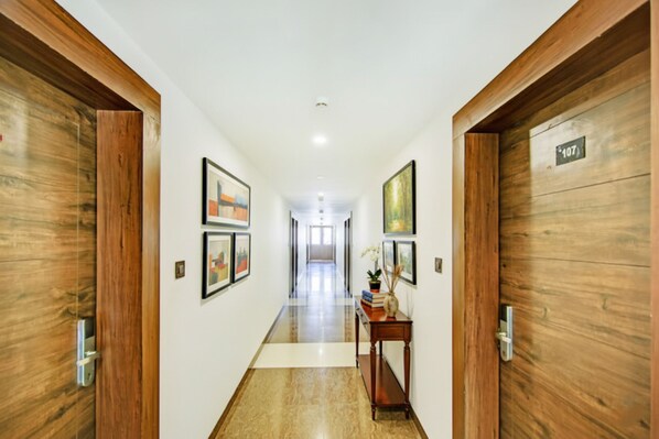 A hallway with wooden doors, framed artwork on the walls, and a small table with books and a vase. - Via Hemkunt Mansion - Nr Noida Sector-15 Metro (Noida)