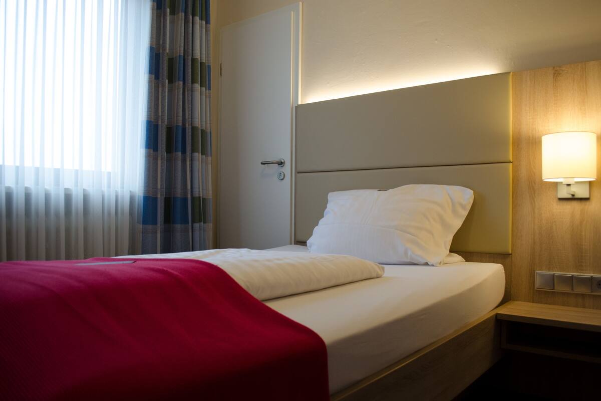 Una habitación de hotel con cama, lámpara de noche y ventana con cortinas.