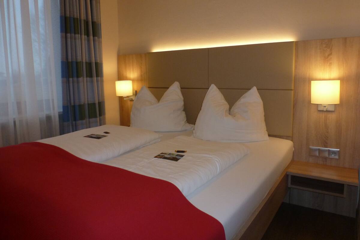 Una habitación de hotel con una cama, dos lámparas de noche, un cabecero de madera y una colcha roja.