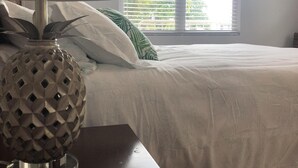 A neatly made bed with white bedding, a lamp with a geometric base, and a window with blinds. - Beautiful Beach Apartment at Sandy Beach, Rincon (Rincon)