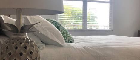 Una cama bien hecha con ropa blanca, una lámpara de base geométrica y una ventana con persianas.