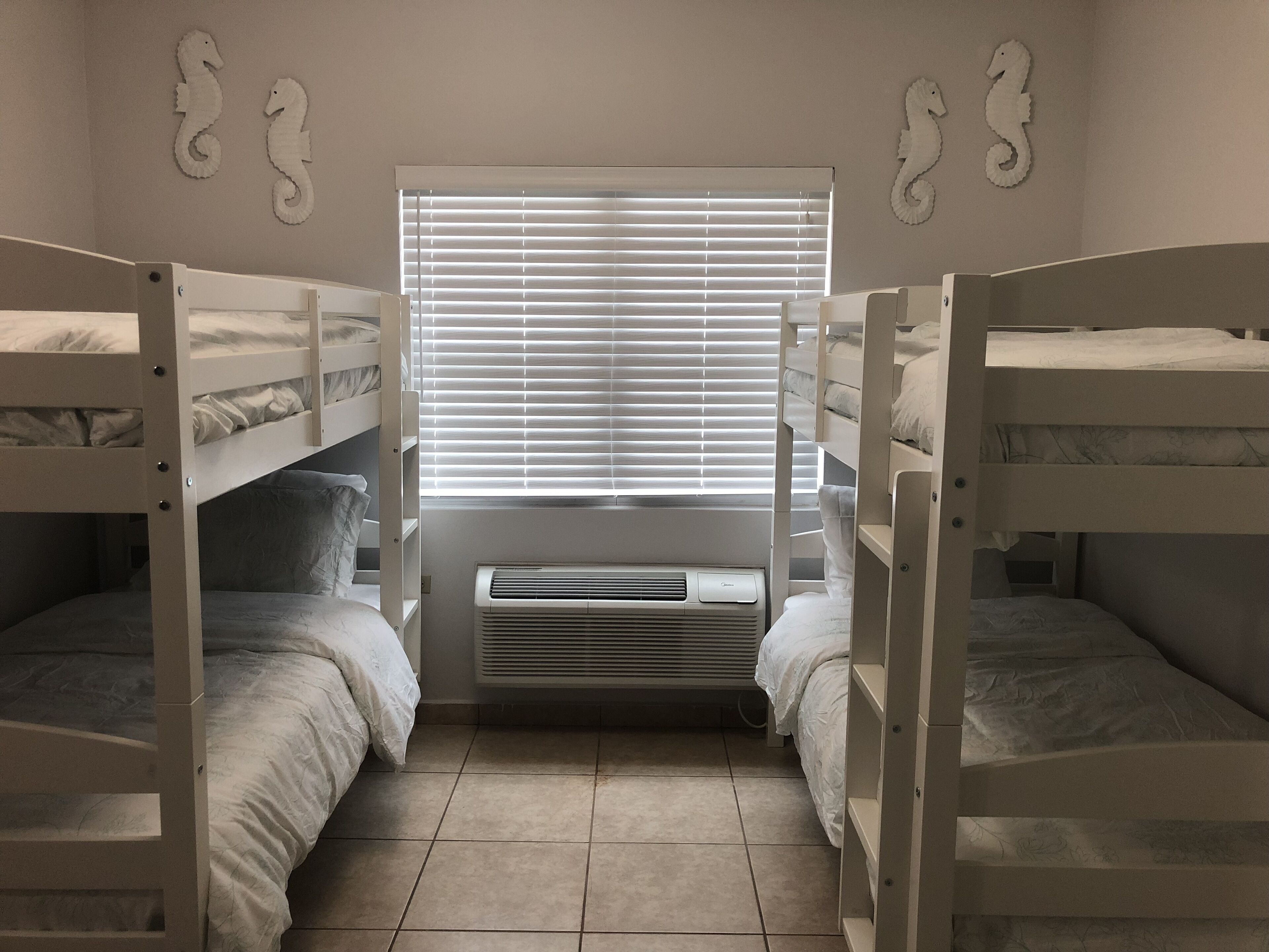 Quarto com beliches, janela com persianas, ar-condicionado fixado na parede e arte decorativa de cavalo-marinho.