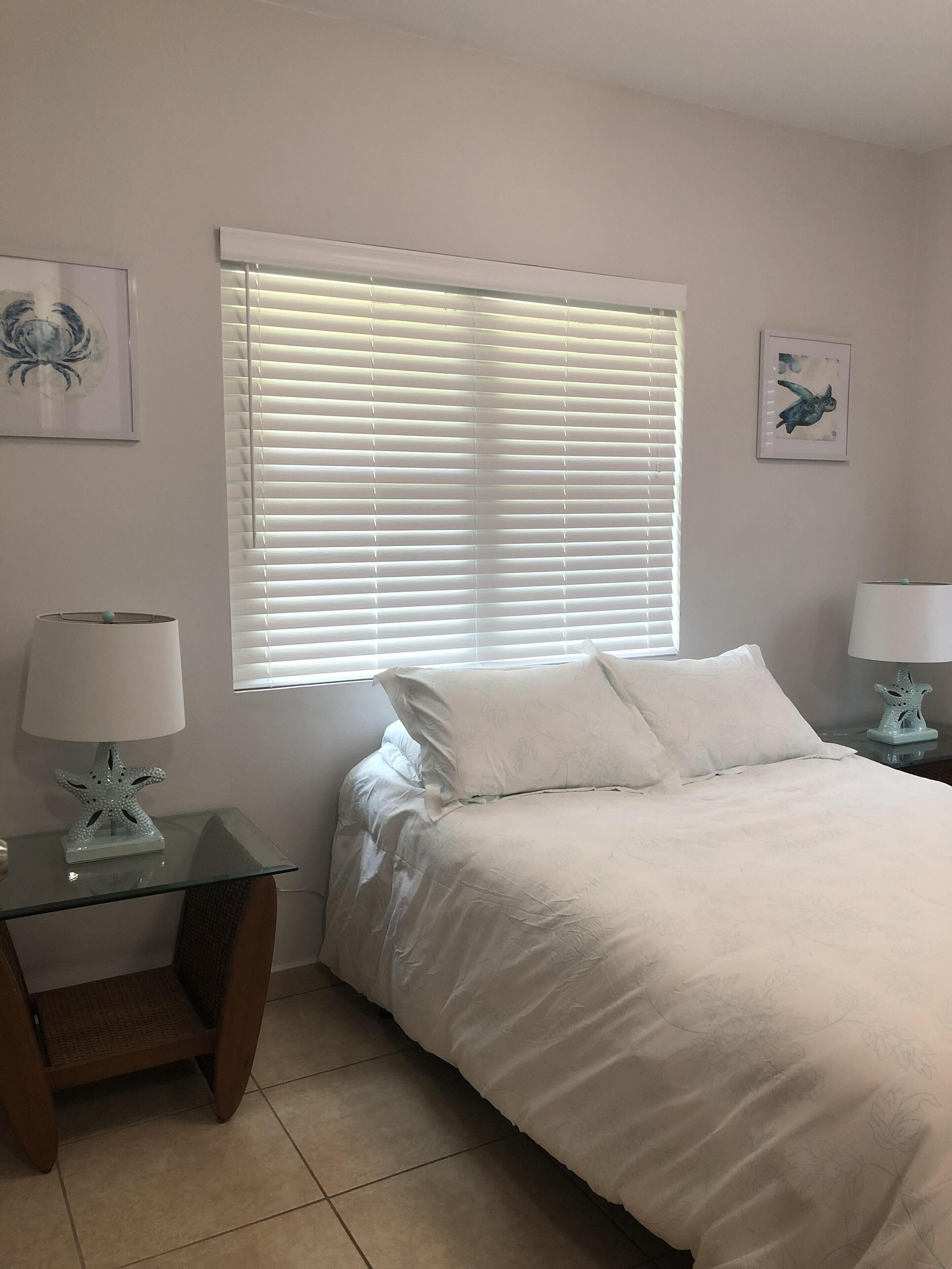 Un dormitorio con cama, mesita de noche, lámpara y ventana con persianas.