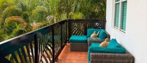 Un balcón con mobiliario de mimbre y cojines color turquesa, rodeado de una barandilla negra, con vistas a un jardín exuberante.