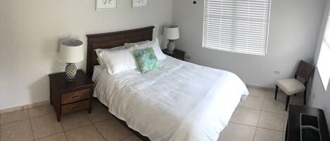 Una cama bien tendida con sábanas blancas, dos mesitas de noche con lámparas, una silla y una obra de arte en la pared.
